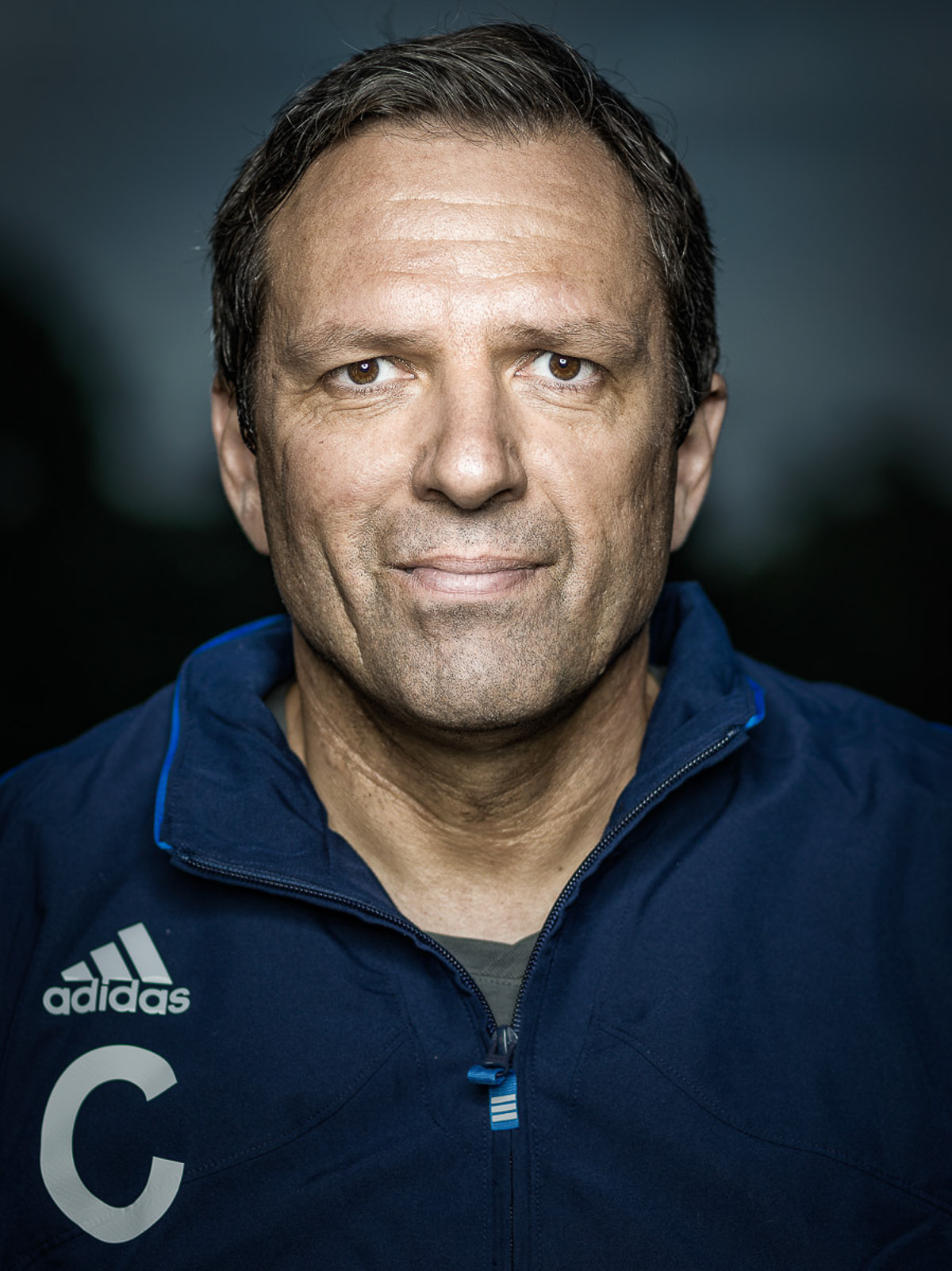 Ein Trainer in blauer Jacke und weißem Hemd steht lächelnd in einem sportlichen Umfeld.