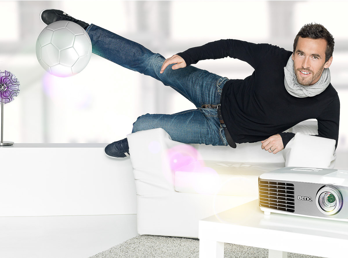 Mann in Jeans und schwarzem Pullover kickt Fußball auf weißem Sofa neben Projektor, modernes Wohnzimmer.