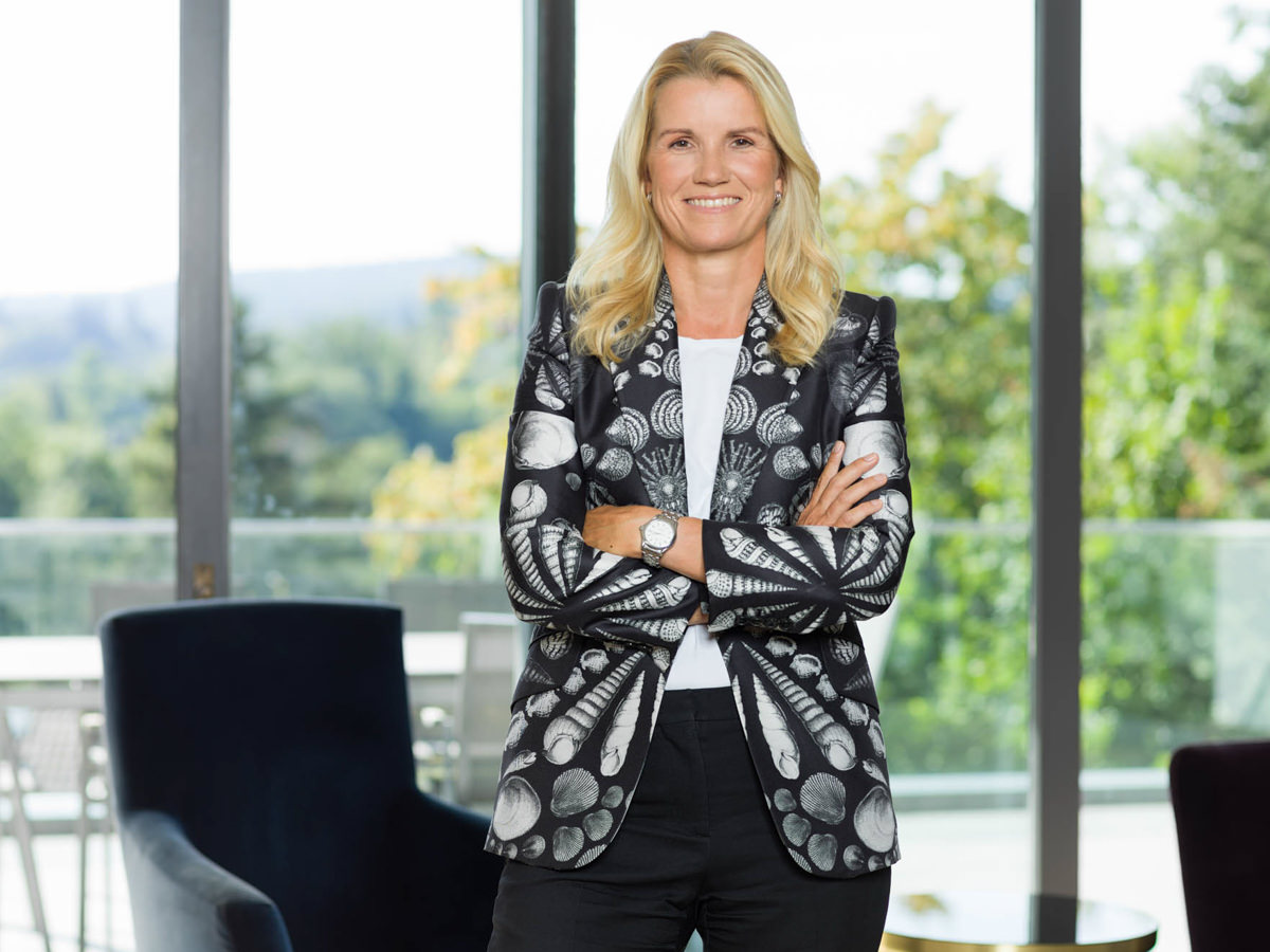 Vorstandsdame im Büro vor Glasfront mit Blick ins Grüne.